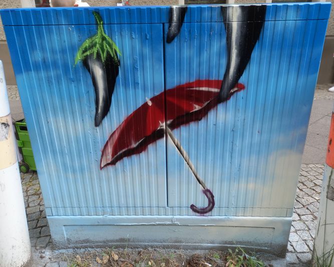 Straßenkunst auf einem Kabelverteiler zeigt drei schwarze Paprikaschoten und einen roten Regenschirm vor einem blauen Himmel mit Wolken.