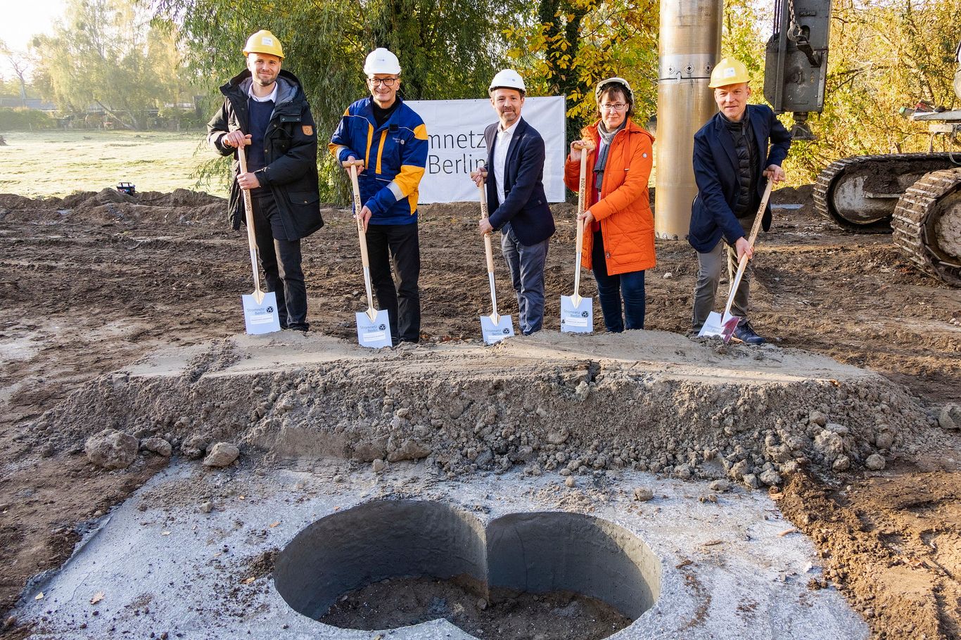 Fünf Menschen mit Bauhelmen halten Schaufeln bei einer Grundsteinlegungszeremonie auf einer Baustelle, im Hintergrund sind ein Banner und Maschinen zu sehen.