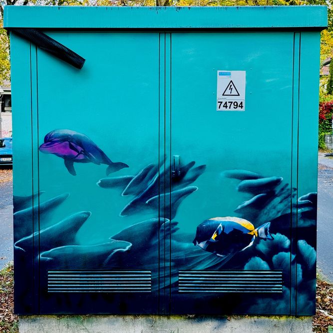 Straßenkunst auf einem Stromkasten zeigt vor einem blaugrünen Hintergrund Unterwasserlebewesen, darunter Fische und einen Delphin.