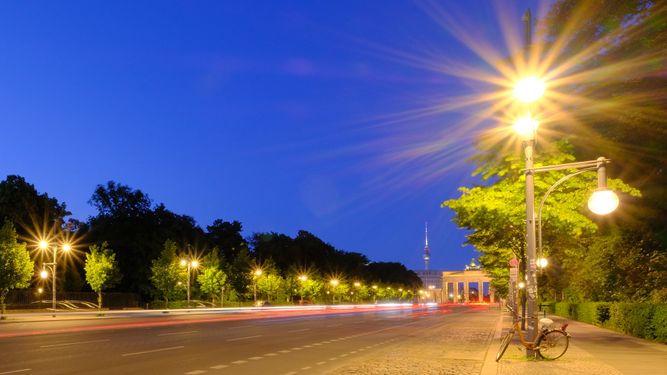 Eine ruhige Stadtstraße, die nachts durch Straßenlaternen beleuchtet wird, mit Bewegungsunschärfe durch vorbeifahrende Fahrzeuge und einem im Hintergrund sichtbaren Wahrzeichen.