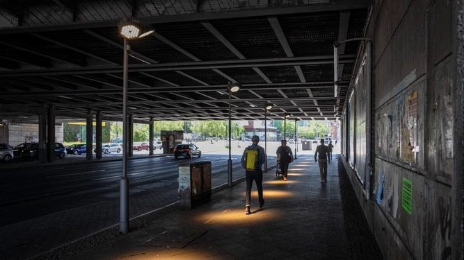 Personen gehen durch eine dunkle Unterführung und auf dem Weg sind helle Lichtpunkte.