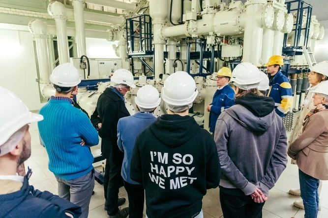 Eine Gruppe von Menschen mit Schutzhelmen versammelt sich um eine Maschinenanlage in einer Industrieumgebung. Einige hören einem Redner in einer blauen Jacke zu.