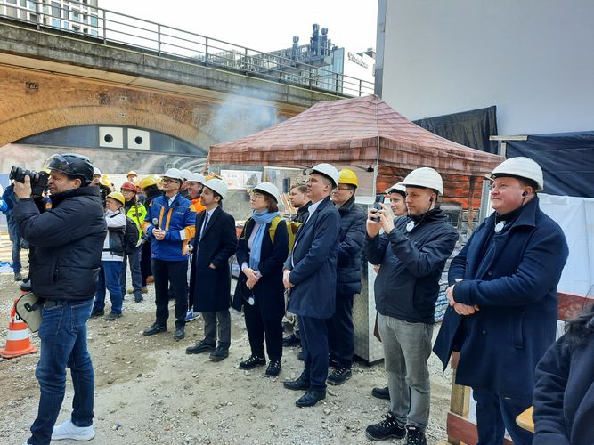 Eine Gruppe von Menschen, meist mit Bauhelmen, besichtigt eine Baustelle. Ein Fotograf hält die Szene fest.