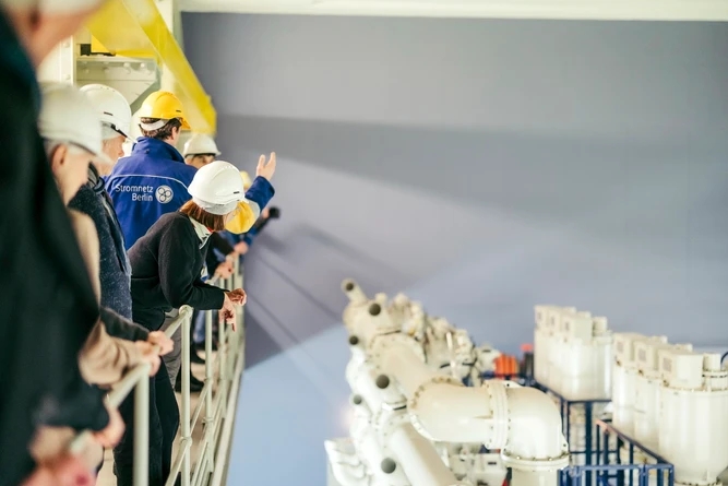 Eine Gruppe von Menschen mit Schutzhelmen hört einem Mann mit gelbem Schutzhelm zu, der in einer Industrieanlage spricht.