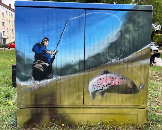 Straßenkunst auf einem Kabelverteiler zeigt eine Person, die tagsüber an einem See angelt, wobei unter einem blauem Himmel ein leuchtender Fisch an der Angelschnur baumelt.