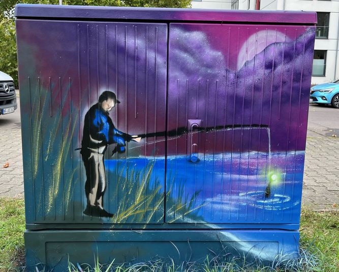 Straßenkunst auf einem Kabelverteiler zeigt eine Person, die nachts an einem See angelt, wobei unter einem mondbeschienenen Himmel ein leuchtender Fisch an der Angelschnur baumelt.