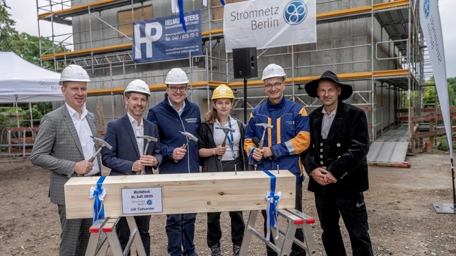 Sechs Personen vor einem Gebäude mit einem Hammer in der Hand und einem Holzbalken im Vordergrund.