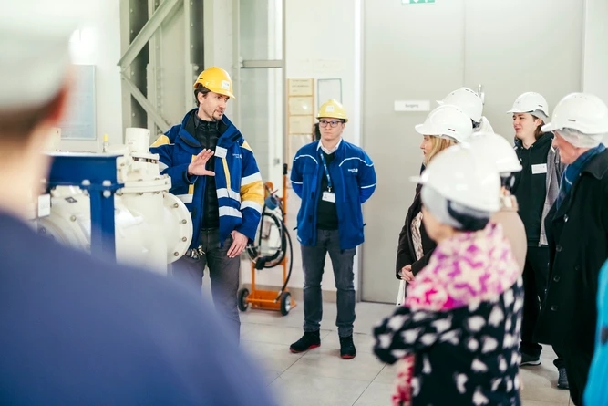 Eine Gruppe von Menschen mit Schutzhelmen hört einer Einweisung in einer Industrieanlage zu.