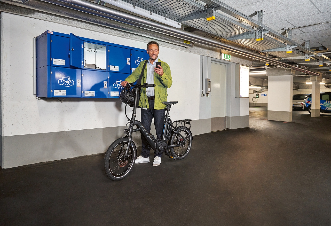 Mann in grüner Jacke und blauer Jeans steht mit seinem Elektro-Fahrrad vor einem blauen Schließfach für Elektro-Fahrrad-Akkus. Die Umgebung zeigt eine gut beleuchtete Tiefgarage.