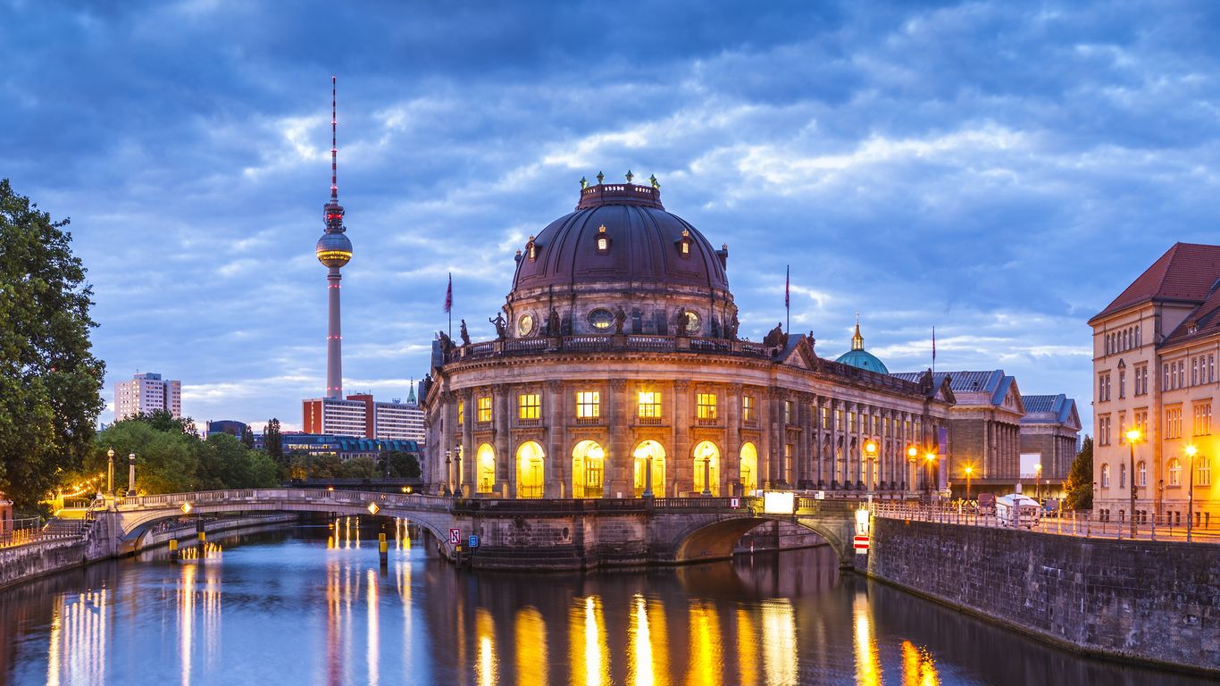 Abendansicht des Berliner Bode-Museums mit dem Fernsehturm im Hintergrund, der sich unter einem bewölkten Himmel in der Spree spiegelt.