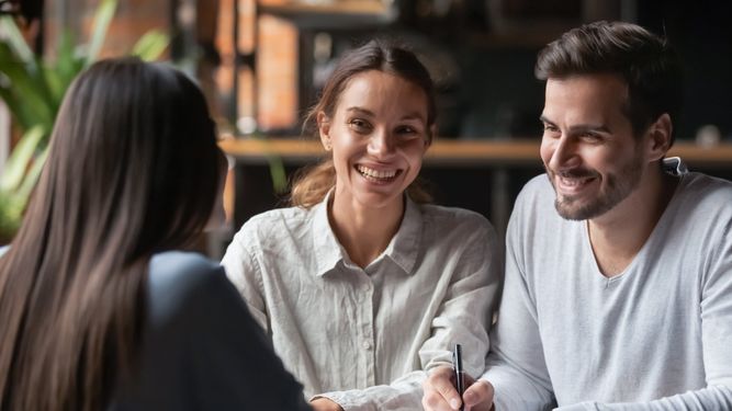 Three people sit at a table; two are smiling and seem engaged in conversation with the third person whose back is to the camera. The setting appears to be a casual indoor environment.