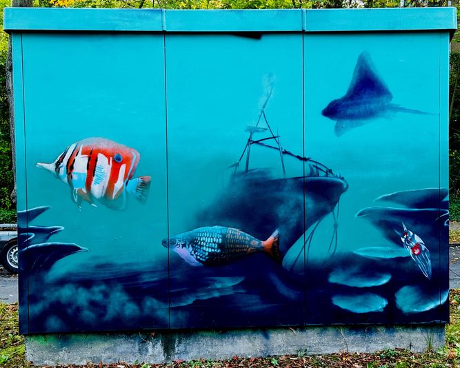 Straßenkunst auf einem Stromkasten zeigt vor einem blaugrünen Hintergrund Unterwasserlebewesen, darunter Fische, einen Stachelrochen und ein gesunkenes Schiff.