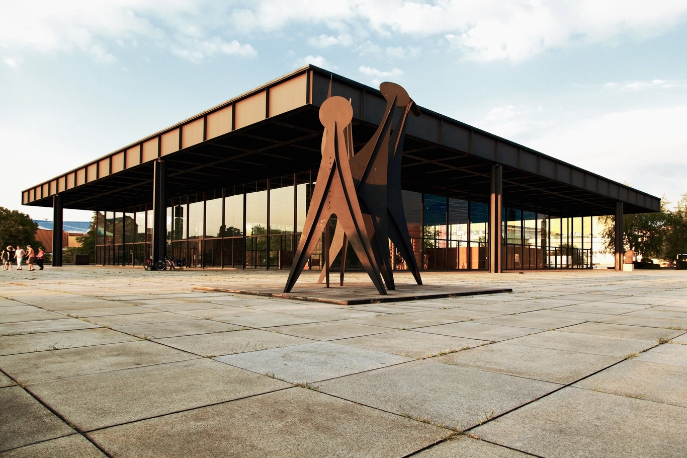 Modernes Gebäude mit weitem Dach und Stützen und davor eine Skulptur unter leicht bewölktem Himmel.
