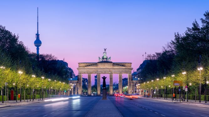 Brandenburg Gate in Berlin at dawn, with the TV Tower in the background. Traffic streaks are visible on the road leading to the gate, lined with illuminated street lamps and trees.
