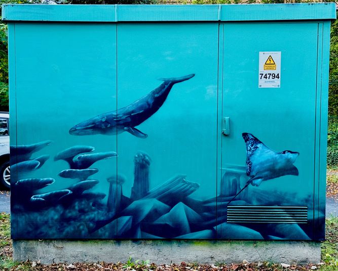 Straßenkunst auf einem Stromkasten zeigt vor einem blaugrünen Hintergrund Unterwasserlebewesen, darunter einen Wal und einen Stachelrochen.
