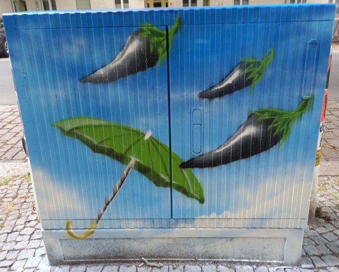 Straßenkunst auf einem Kabelverteiler zeigt drei schwarze Paprikaschoten und einen grünen Regenschirm vor einem blauen Himmel mit Wolken.