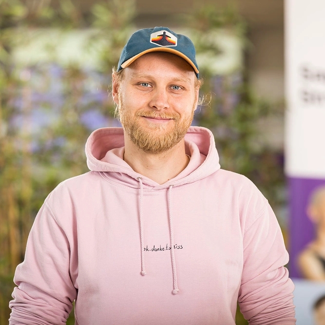 Lächelnder Mann mit Bart, hellen Haaren, dunkelblauer Kappe mit Berg-Patch und rosa Hoodie.